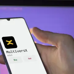 Hand hält Smartphone mit MultiversX App, die EGLD-Token anzeigt. Kauf- und Verkaufsoptionen sind sichtbar.