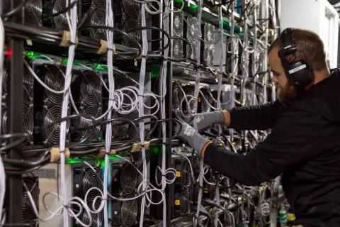 Ein Mann mit Kopfhörern arbeitet an einer Wand voller Lüfter und Kabel. Dies zeigt, wie funktioniert krypto mining: leistungsstarke Computer lösen komplexe Rätsel.