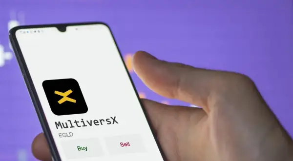 Hand hält Smartphone mit MultiversX App, die EGLD-Token anzeigt. Kauf- und Verkaufsoptionen sind sichtbar.