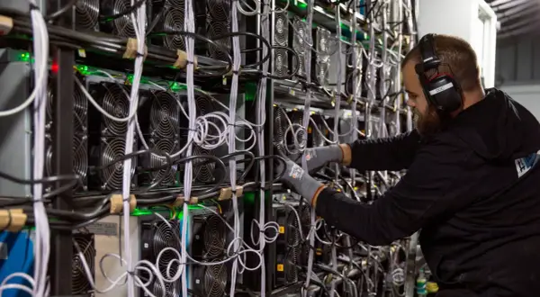 Ein Mann mit Kopfhörern arbeitet an einer Wand voller Lüfter und Kabel. Dies zeigt, wie funktioniert krypto mining: leistungsstarke Computer lösen komplexe Rätsel.