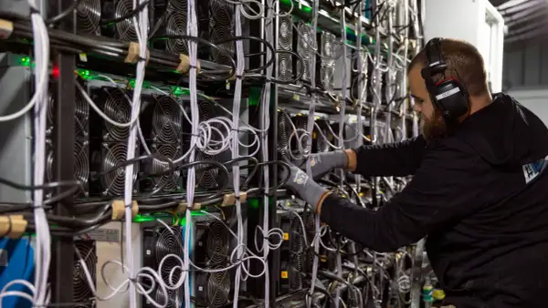 Ein Mann mit Kopfhörern arbeitet an einer Wand voller Lüfter und Kabel. Dies zeigt, wie funktioniert krypto mining: leistungsstarke Computer lösen komplexe Rätsel.